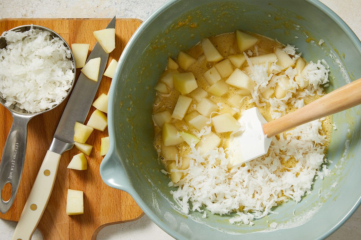 Overhead shot of diced pears being stirred into the batter in a gray bowl with a rubber spatula, wooden cutting board and knife beside it.