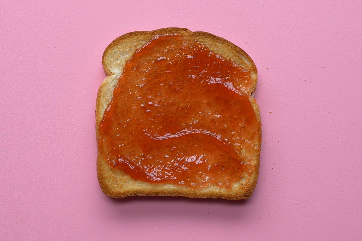 This is a close-up shot of toasted bread with orange-red fruit jam on top; the background is pink