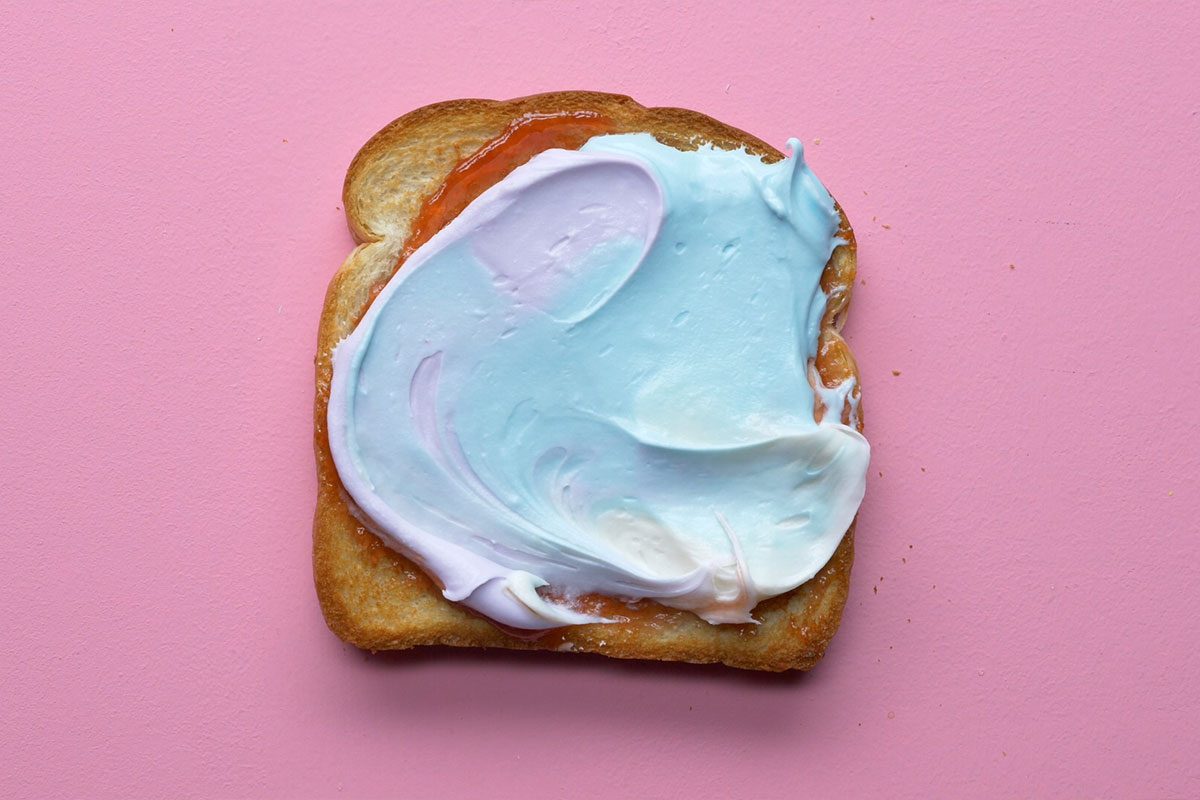 This is a close-up shot: A slice of toast with pastel blue; pink; and white swirled spread sits on a pink surface