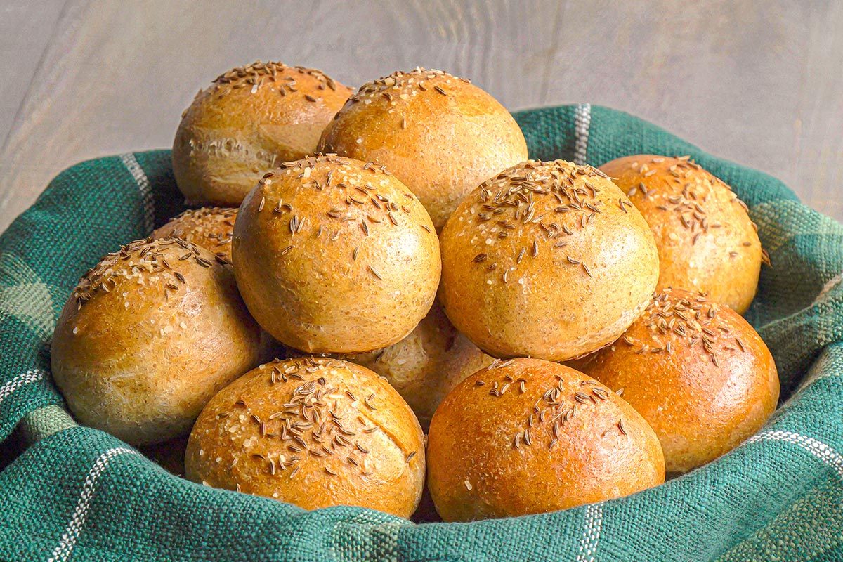 Closeup shot of a basket lined with green checkered cloth holding a pile of rye rolls round, golden brown, and topped with caraway seeds;