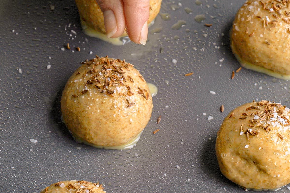 Closeup shot of a hand sprinkling seeds over round dough balls arranged on a greased baking tray, preparing them for baking;