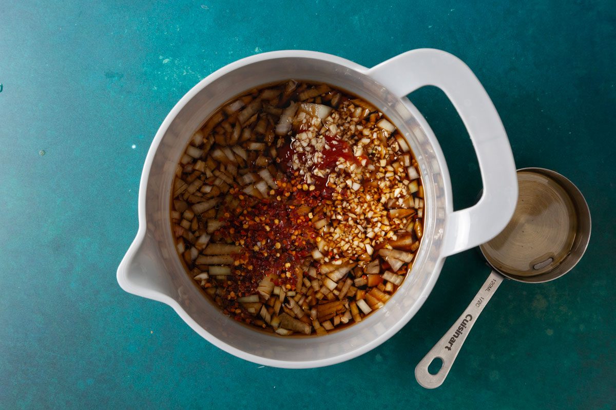 Overhead shot of a white mixing bowl filled with diced onions, minced garlic, crushed red pepper flakes, and sauces being combined into a marinade; a metal measuring cup rests beside it on a teal surface.