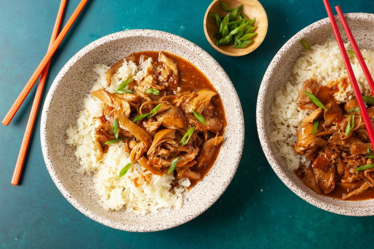 Overhead shot of two speckled bowls filled with white rice topped with tender shredded honey-garlic chicken in a glossy brown sauce, garnished with sliced green onions; chopsticks rest beside the bowls on a teal surface;