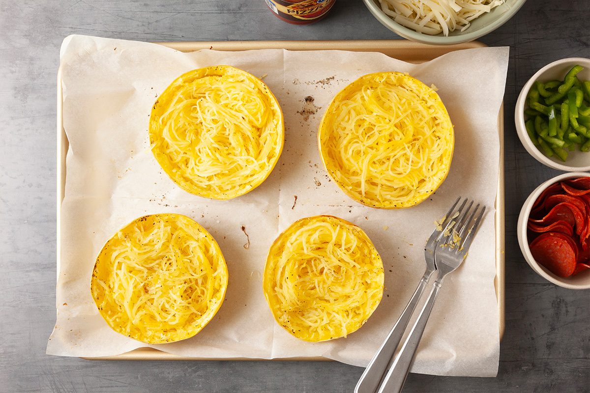 Overhead shot of a parchment-lined baking sheet with four roasted squash halves filled with fork-shredded strands; A fork rests beside them, and small bowls of sliced peppers, pepperoni, and shredded cheese sit nearby on the gray surface;