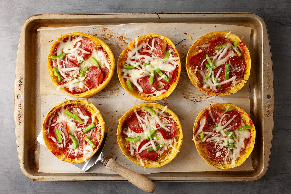 Overhead shot of a metal baking sheet with six baked spaghetti squash pizzas topped with melted cheese, pepperoni, and sliced green peppers; A small spatula slides under one pizza, revealing the golden squash edges;