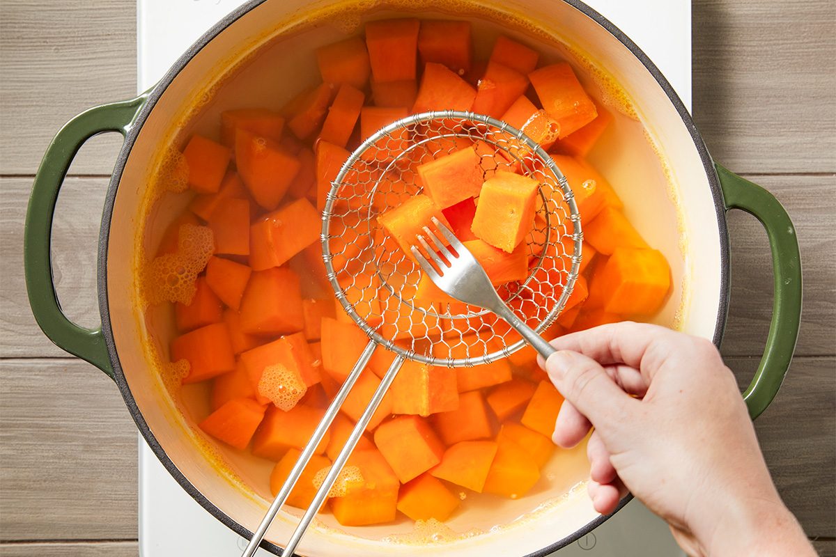 Overhead shot of a hand using a slotted spoon and fork to lift cooked orange sweet potato cubes from a pot of hot water on the stove;