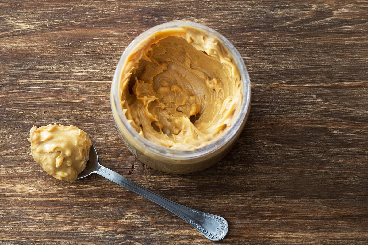 A jar of chunky peanut butter sits on a wooden surface, with a metal spoon holding a scoop of the peanut butter placed beside it.