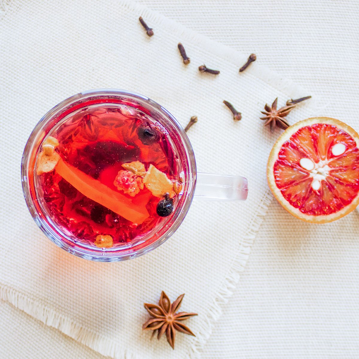 A glass cup of red herbal tea with visible fruit and spices, placed on a white cloth next to a halved blood orange, star anise, and cloves.