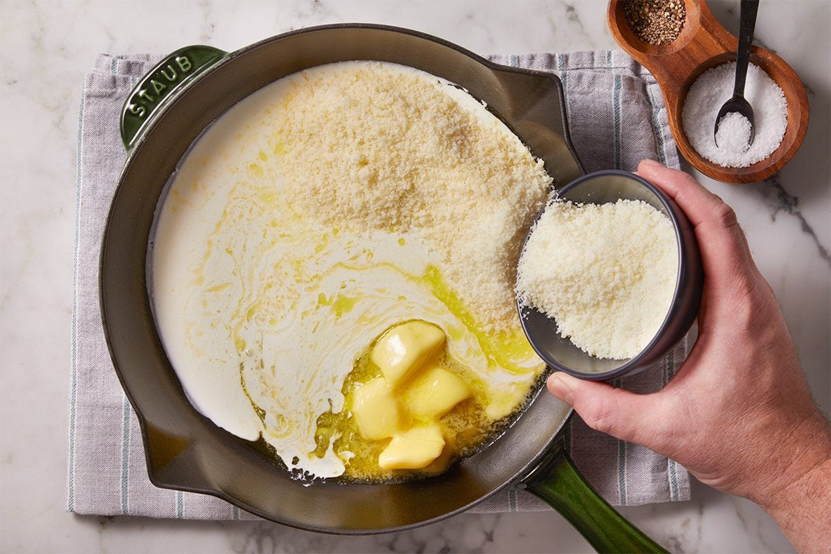 Overhead shot of a hand adds grated cheese to a skillet with melted butter and milk the pan rests on a gray towel near salt and pepper bowls