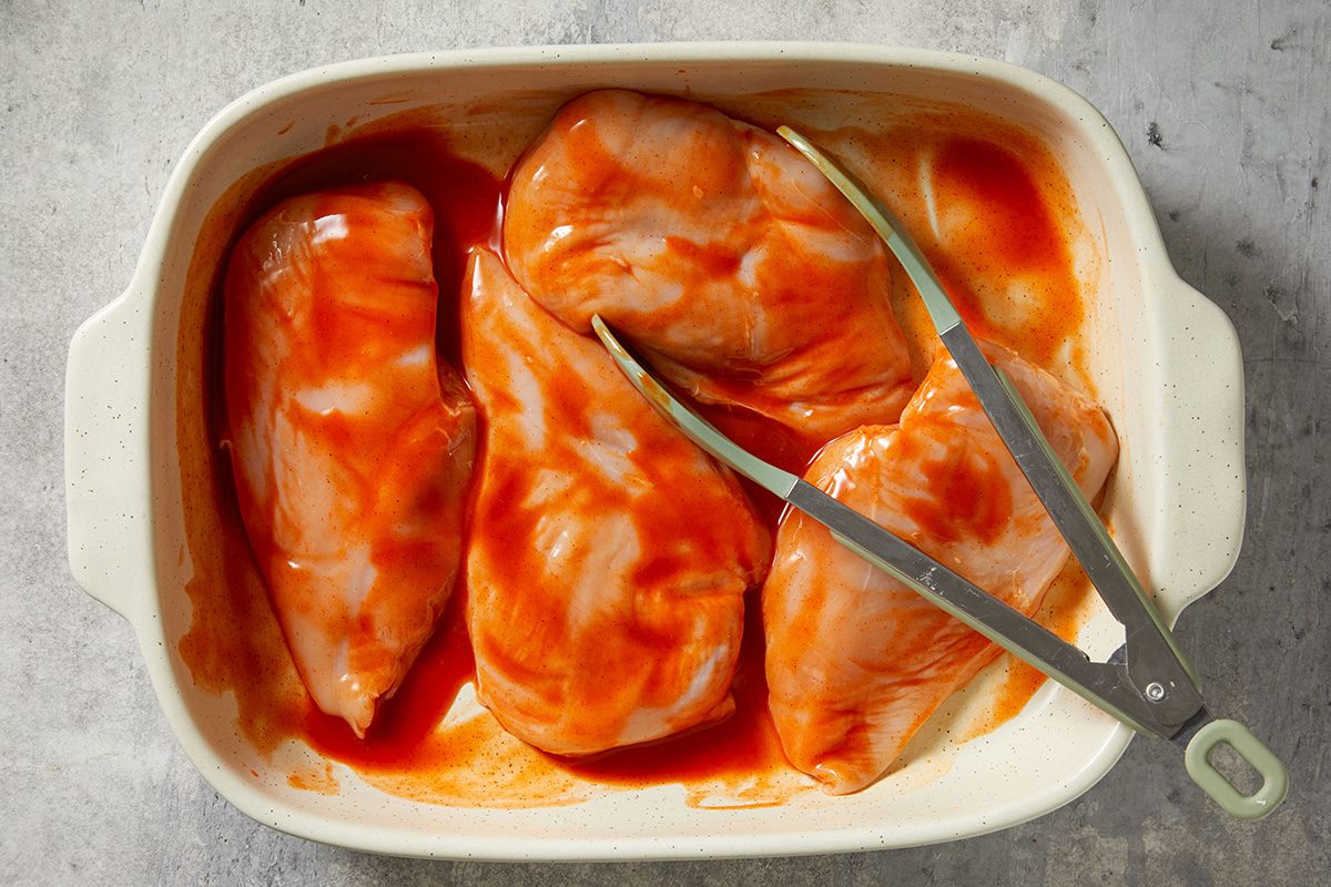 Four raw chicken breasts coated in red marinade sit in a white baking dish. Metal tongs rest on top of one chicken breast. The background surface is light gray.