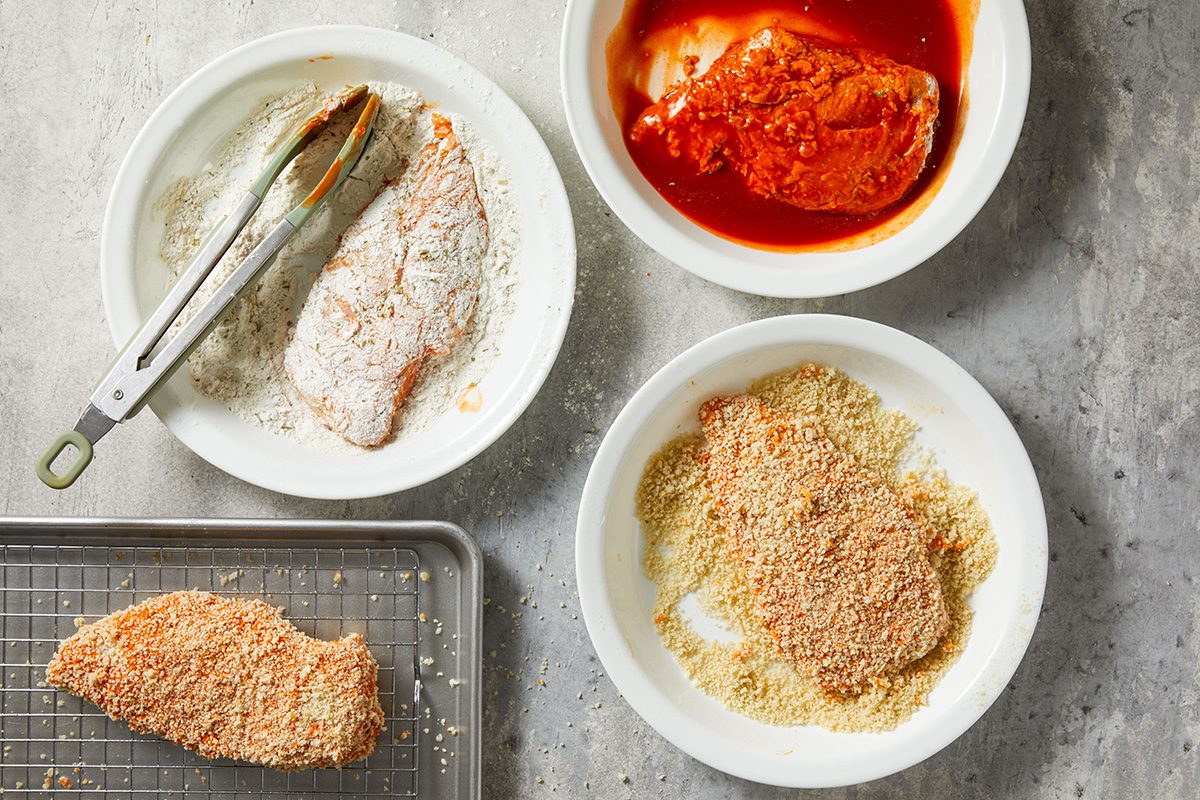 Four bowls show the steps of breading chicken: one with flour, one with red sauce, one with breadcrumbs, and one finished breaded chicken breast on a rack. Metal tongs hold a chicken piece in the flour bowl.