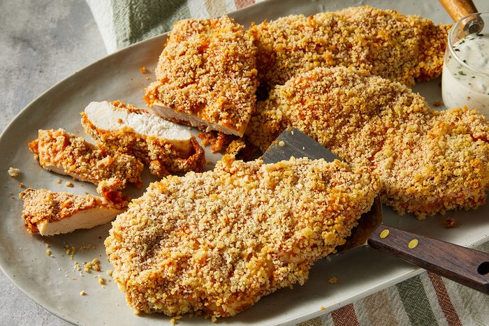 A platter of golden, crispy breaded chicken cutlets, some sliced, with a serving utensil and a small bowl of dipping sauce nearby on a striped towel.