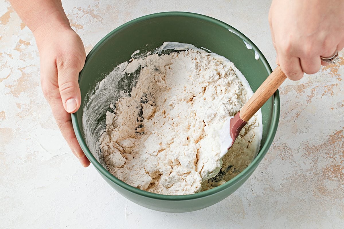 Overhead shot of a pair of hands carefully blends flour and other visible ingredients inside a green bowl using a spatula the bowl sits on a pale countertop