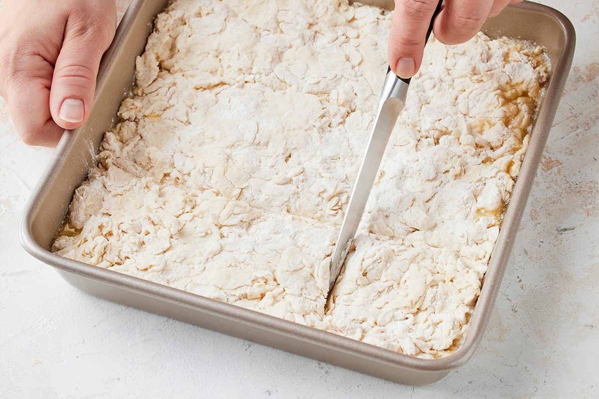 3/4th shot of a person with visible hands holds a shiny knife slicing pale flourdusted dough into even squares inside a rectangular metal baking pan