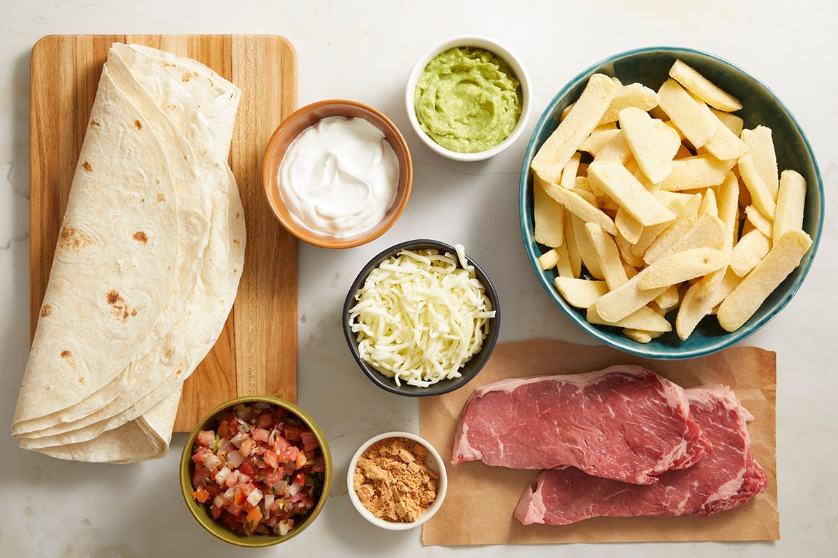 Ingredients laid out on a white surface: flour tortillas, raw steak, shredded cabbage, guacamole, sour cream, salsa, shredded cheese, and a bowl of uncooked fries.