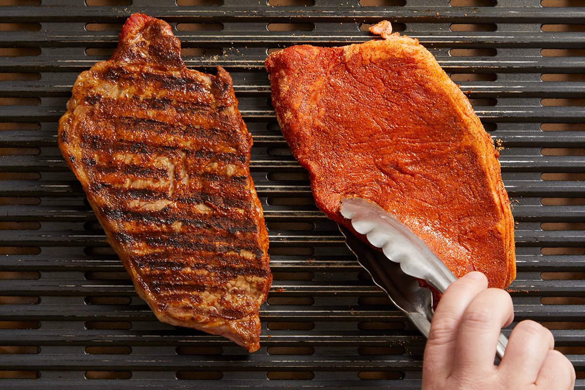 Two steaks on a grill, one with grill marks and one being placed down with metal tongs by a hand.