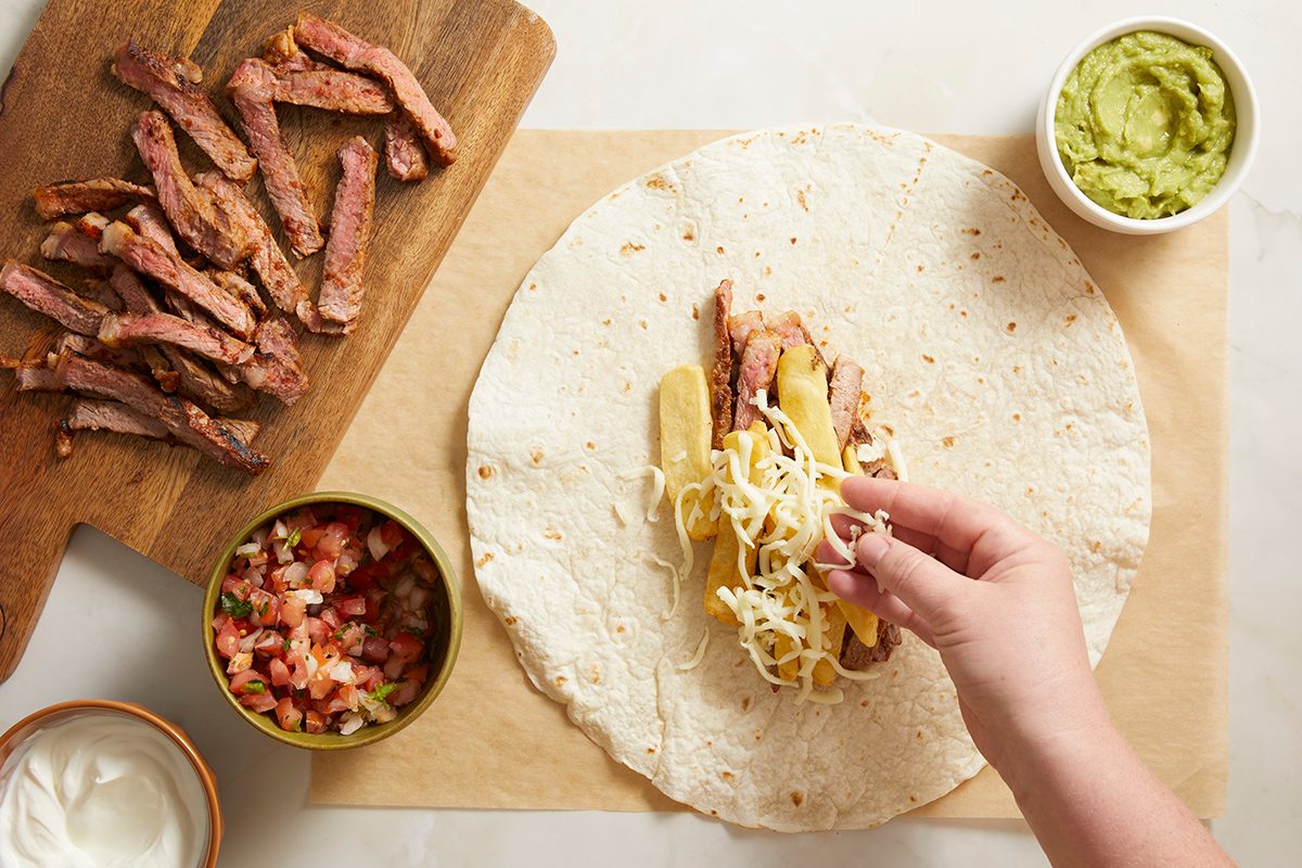 A hand adds shredded cheese to a tortilla filled with sliced steak and vegetables. Nearby are bowls of guacamole, salsa, and a wooden board with more steak strips.