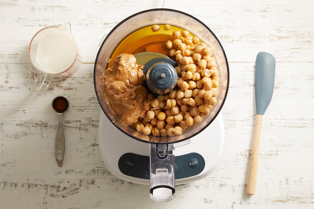 Top-down view of a food processor containing chickpeas, peanut butter, honey, and vanilla. Beside it are a rubber spatula, a spoon with cocoa powder, and a cup of milk on a white wooden surface.