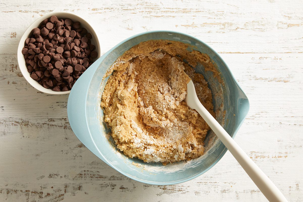 A blue bowl with cookie dough and a white spatula sits beside a small white bowl filled with chocolate chips on a white wooden surface.