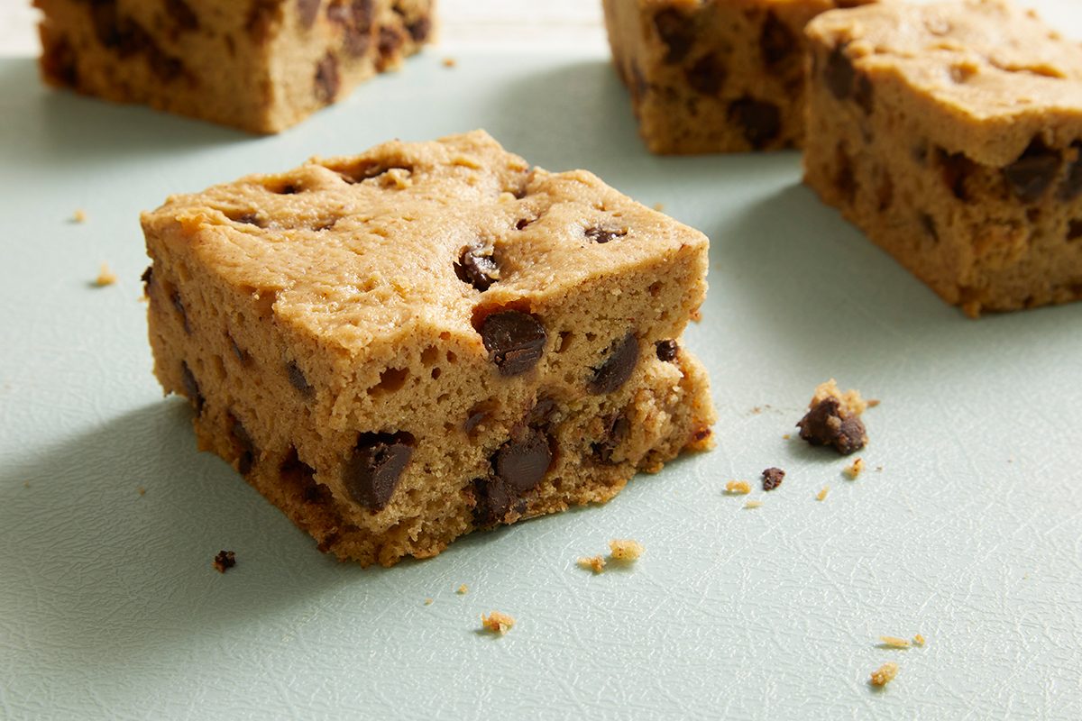 A close-up of thick, square chocolate chip cookie bars with a golden-brown color, resting on a light blue surface. Chocolate chips are visible throughout each bar.