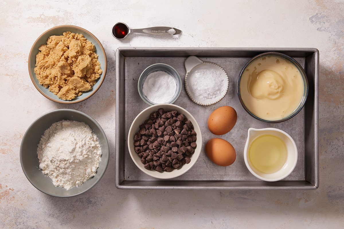 A tray contains eggs, oil, chocolate chips, vanilla pudding, powdered sugar, and baking soda. Nearby are bowls of flour and brown sugar, plus a teaspoon of vanilla extract. All are arranged on a light countertop.