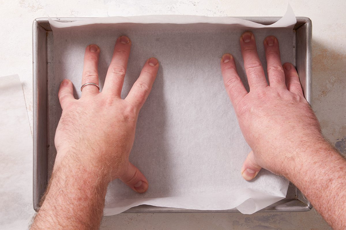 Two hands pressing parchment paper into the bottom of a rectangular baking pan, preparing it for baking.