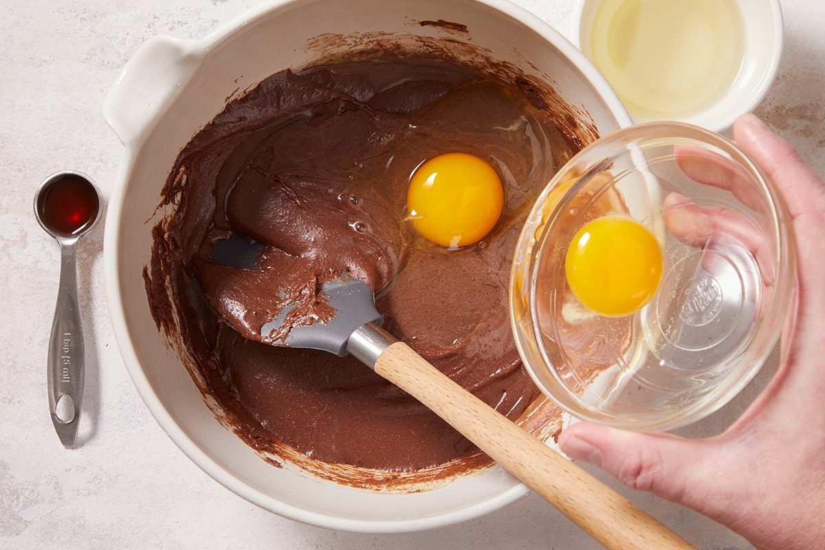 A bowl of chocolate batter with two eggs on top, a hand holding a small bowl near the eggs, a spatula resting in the batter, and measuring spoons and a cup of oil nearby.