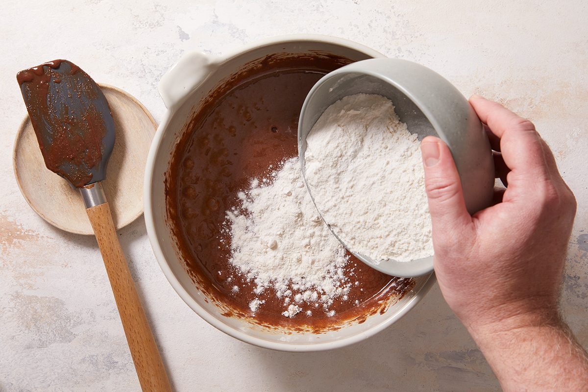 A hand is pouring flour from a bowl into a larger bowl of chocolate batter. A spatula with chocolate residue rests on a small plate nearby on a light-colored surface.