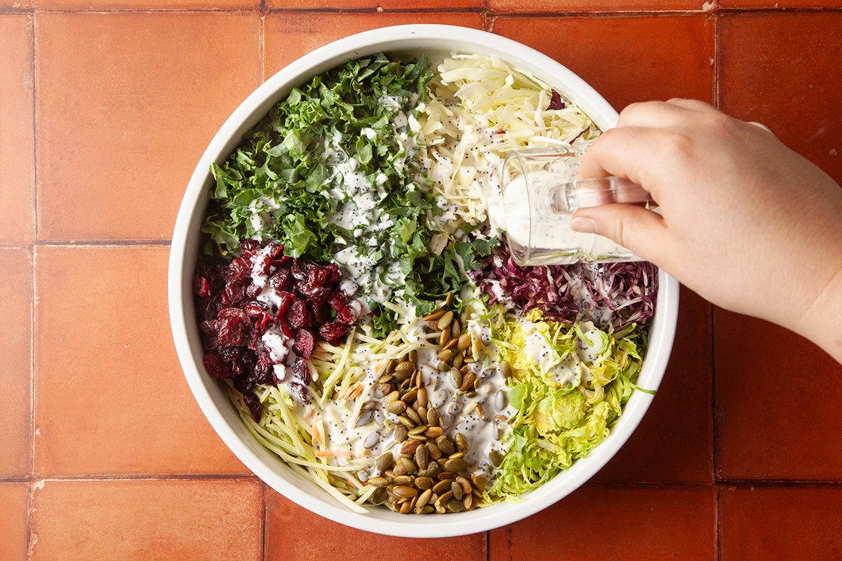 Overhead view of a large bowl filled with chopped kale, shredded cabbage, dried cranberries, and seeds, with a hand pouring creamy dressing over the salad.