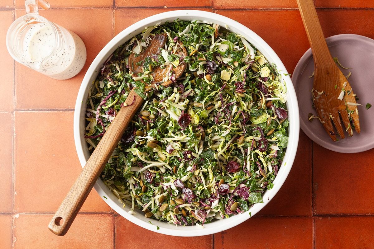 Overhead shot of sweet kale salad being tossed with wooden utensils in a large bowl, showing greens evenly coated with dressing.
