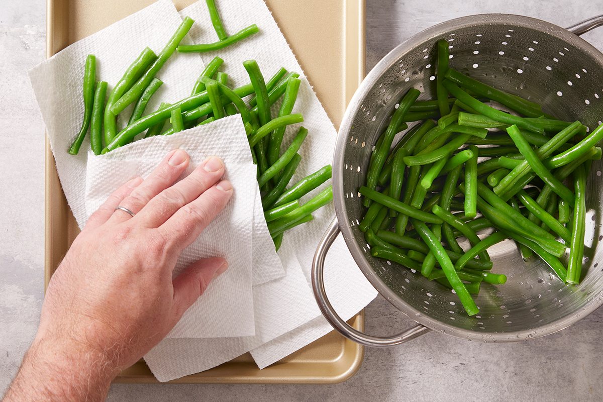 A hand patting green beans dry with a paper towel on a tray, while more green beans are in a metal colander nearby.