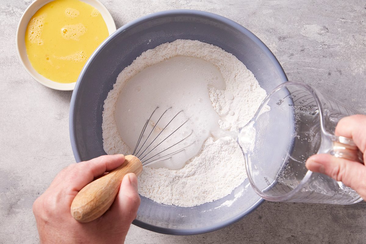 A person pours liquid from a measuring cup into a bowl of flour and sugar while holding a whisk, with a bowl of beaten eggs nearby on a gray countertop.