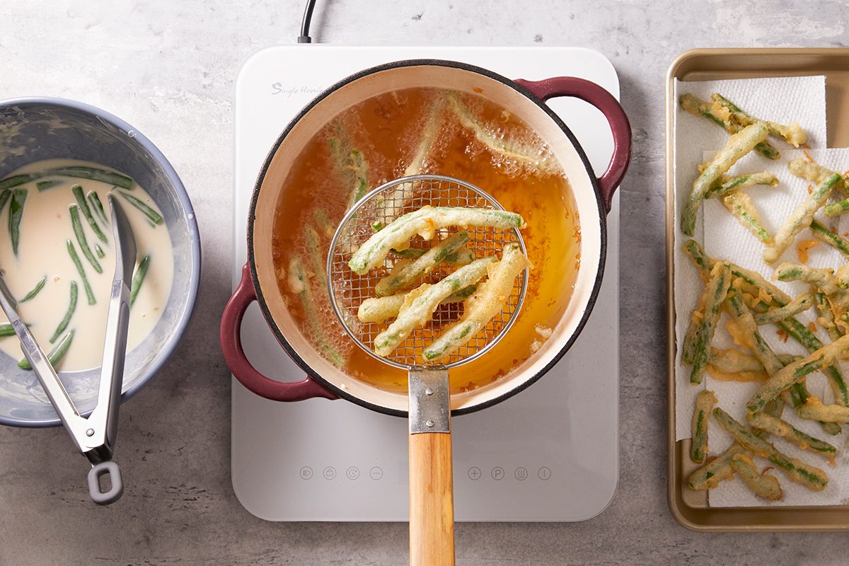 A pot of oil sits on a stovetop with battered vegetables being fried; some are on a slotted ladle above the oil. To the left is a bowl of batter with tongs, and to the right, fried vegetables on a tray.