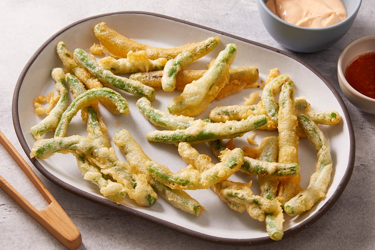 A plate of crispy tempura green beans and yellow beans is served with a side of dipping sauces, one creamy and one red, with wooden tongs resting nearby.