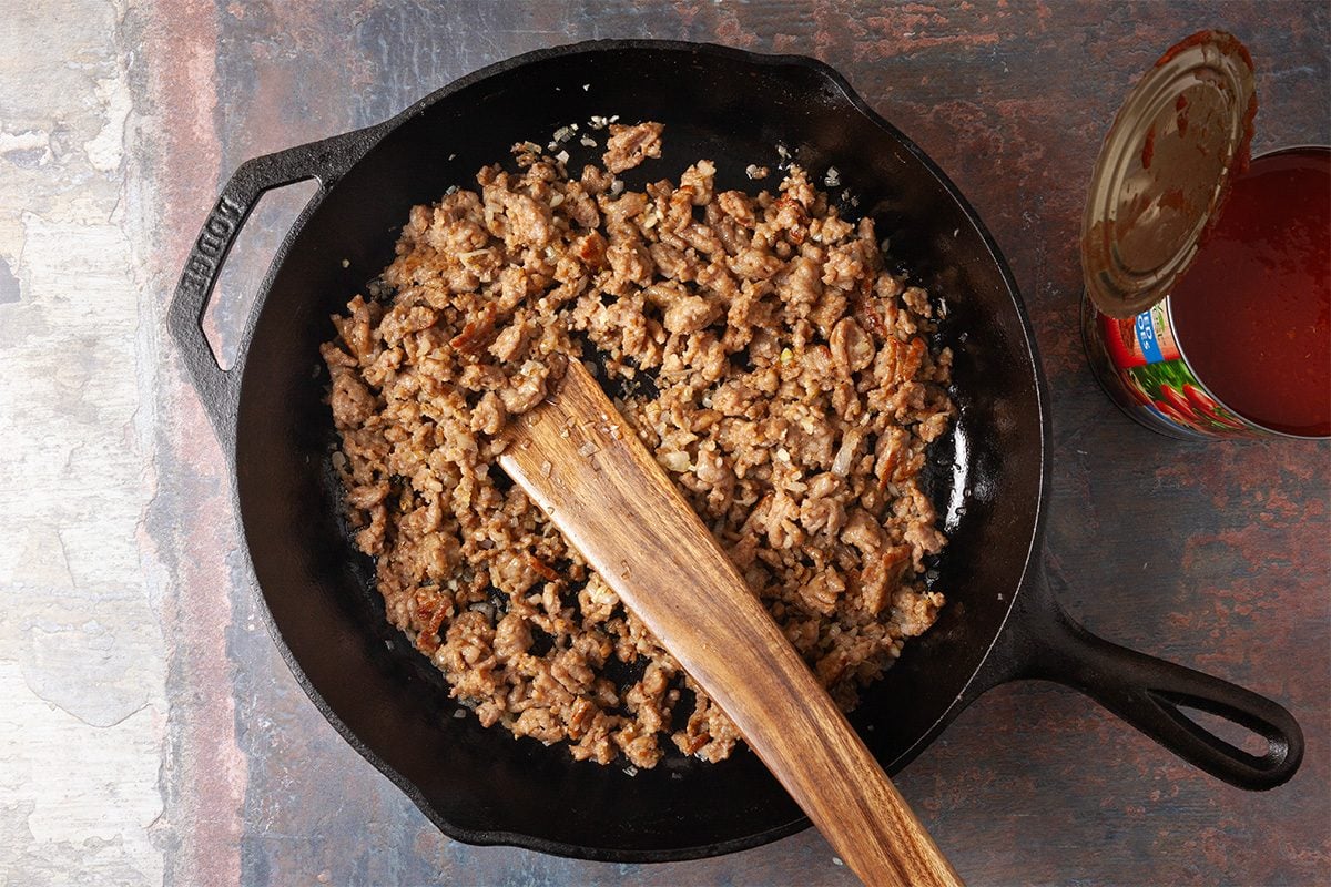 Overhead, horizontal step-by-step image of bulk Italian sausage browning in a cast iron skillet with olive oil. A wooden spoon stirs the sausage as it cooks, forming the savory base for a gnocchi lasagna bake recipe.