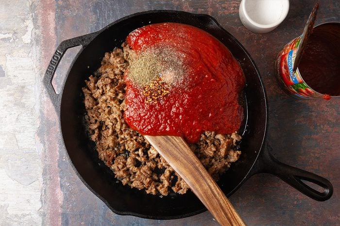 Overhead, horizontal step-by-step image of crushed tomatoes and seasonings being added to browned Italian sausage in a cast iron skillet. The tomato sauce begins forming the base of a gnocchi lasagna bake.