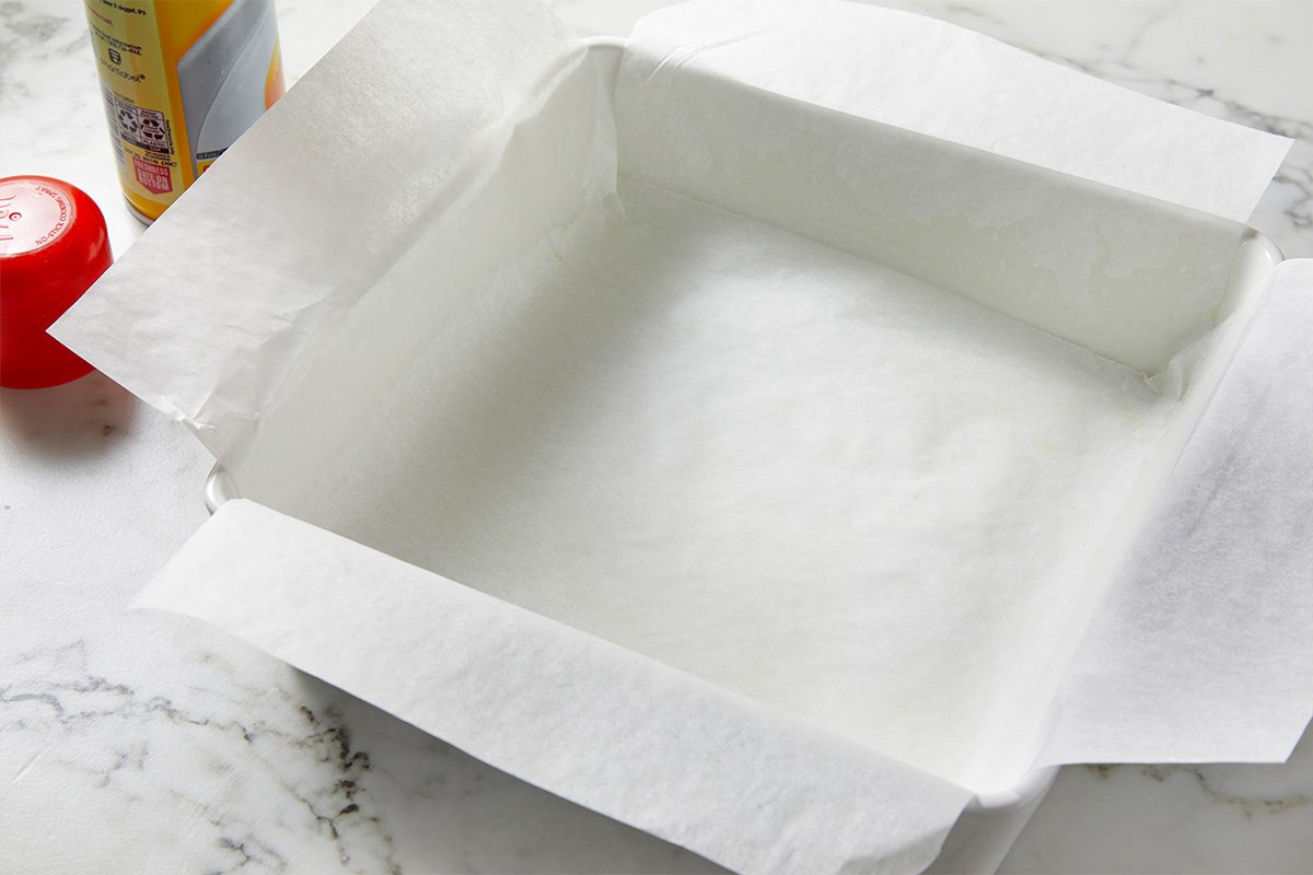 Overhead shot of a square baking pan lined with parchment paper on a marble countertop, with a can of nonstick cooking spray and a red measuring cup partially visible beside it;