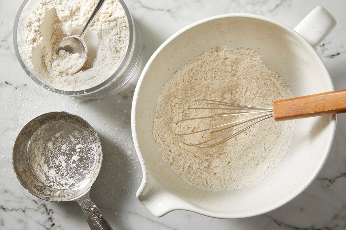 Overhead shot of a white mixing bowl with flour and a whisk, next to a metal measuring cup of flour and a glass jar of flour on a marble countertop;