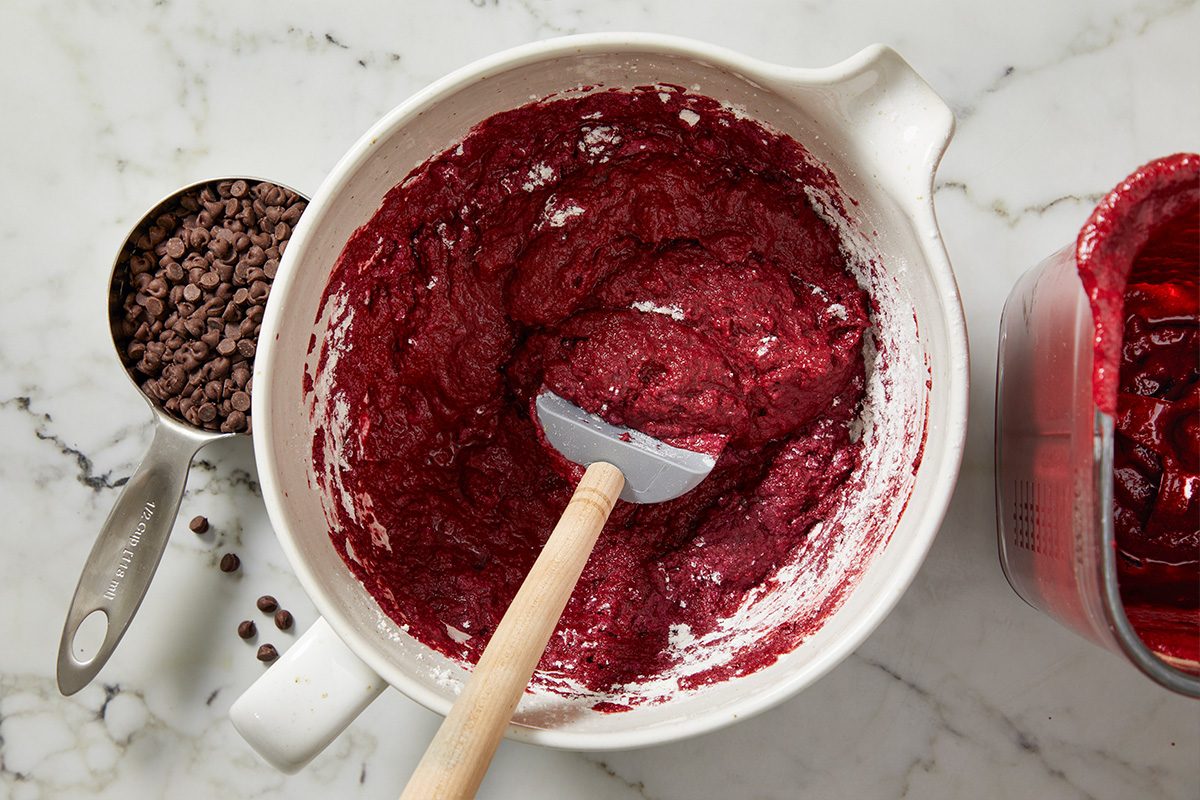 Overhead shot of a white mixing bowl with red batter and a spatula on a marble countertop, next to a metal measuring cup of chocolate chips and a container of red mixture;