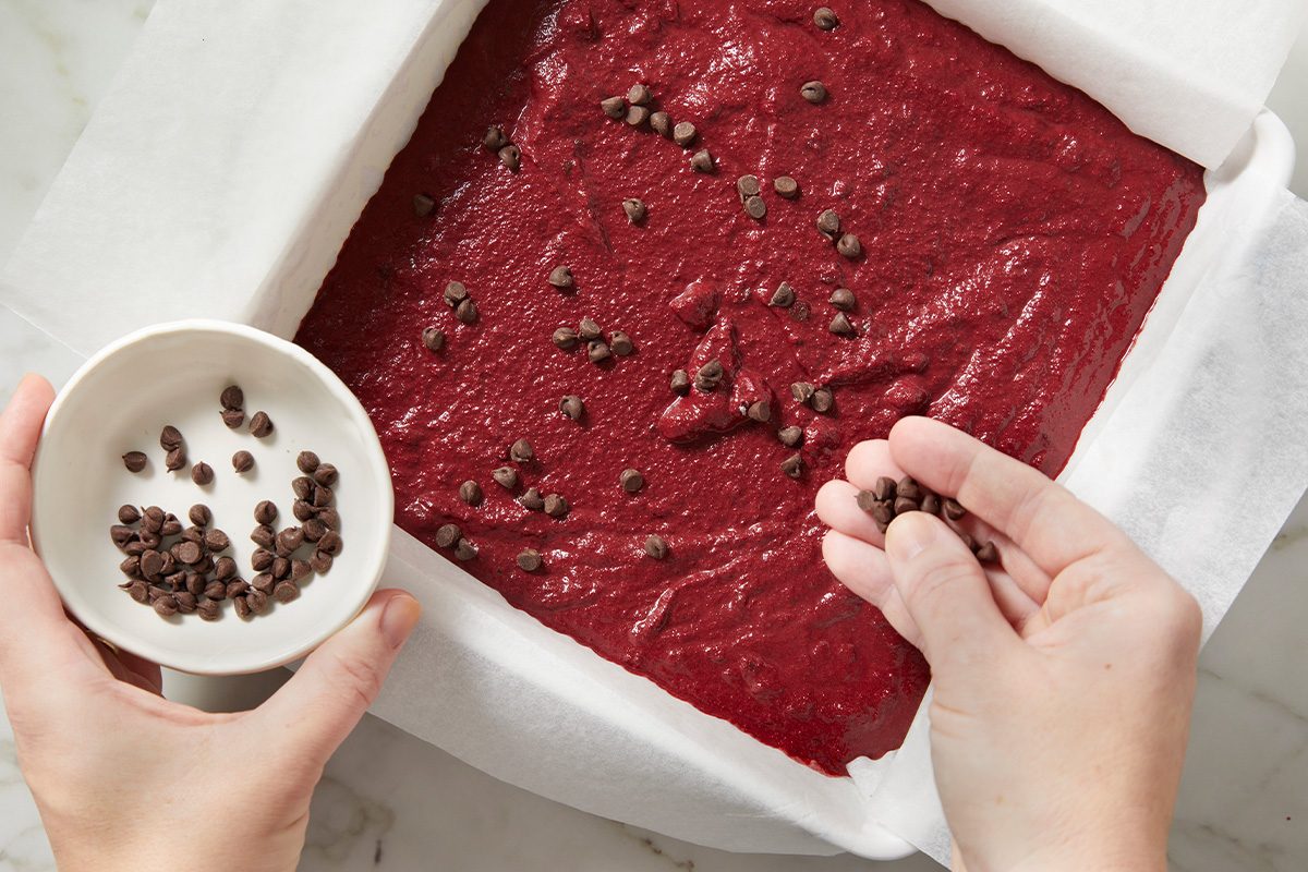 Overhead shot of a person sprinkling chocolate chips from a small bowl onto a pan of red batter lined with parchment paper;