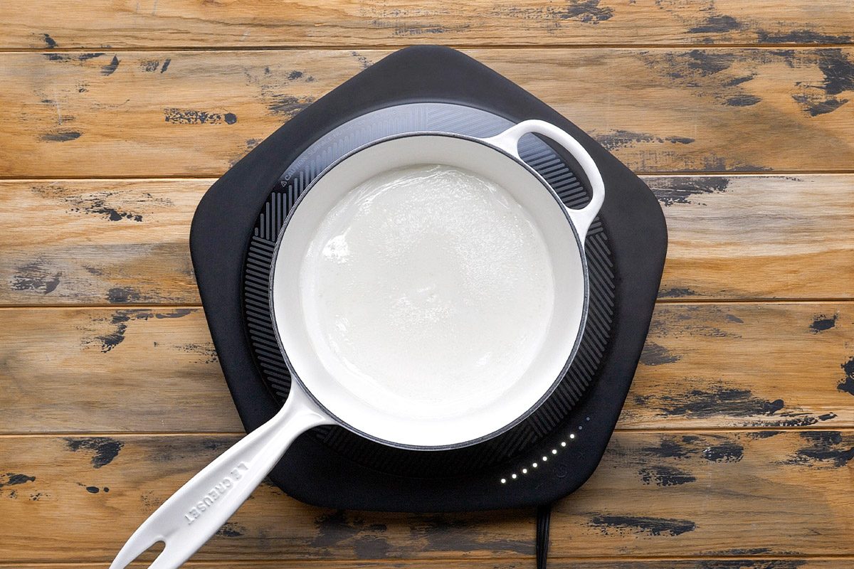 Overhead shot of a white saucepan with a handle is on a black electric cooktop and heats a clear liquid and the cooktop rests on a rustic wooden table