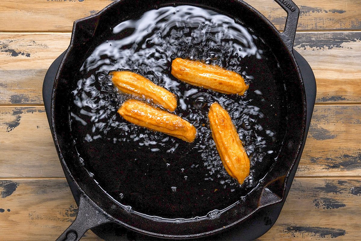 Overhead shot of a Four pieces of dough sizzle in bubbling oil inside a black cast iron skillet the pan rests on a rustic wooden surface the heat is visible