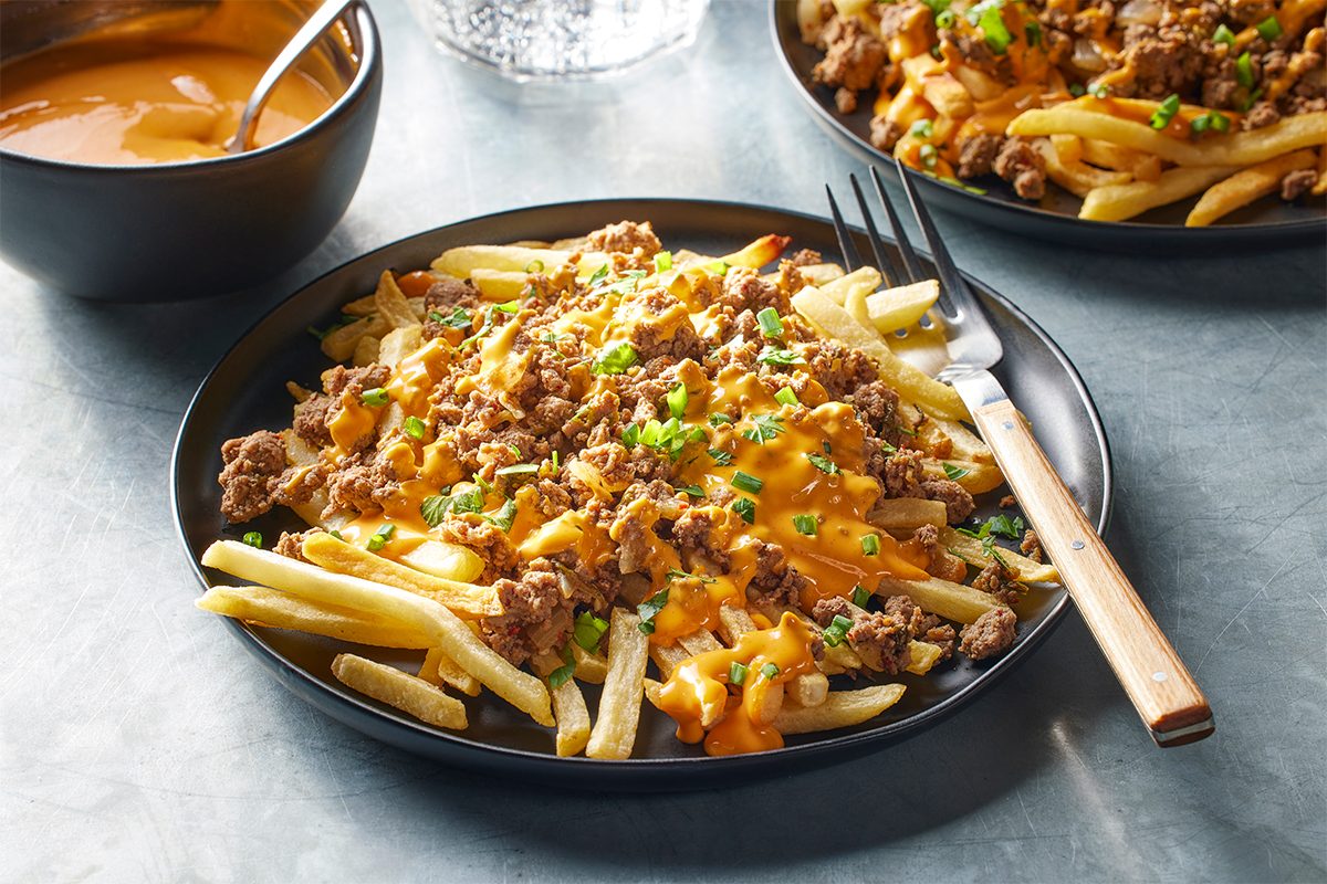 A black plate of French fries is topped with melted cheese, seasoned ground beef, and chopped green onions. A fork rests on the plate, with a bowl of cheese sauce and another plate of the same dish in the background.