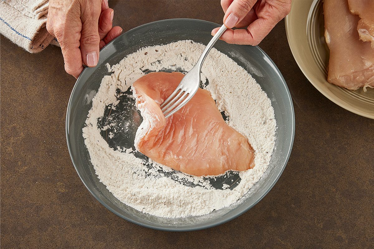 Overhead shot of a person uses a fork to dip a raw chicken breast into flour on a gray plate nearby more chicken breasts rest on another plate;
