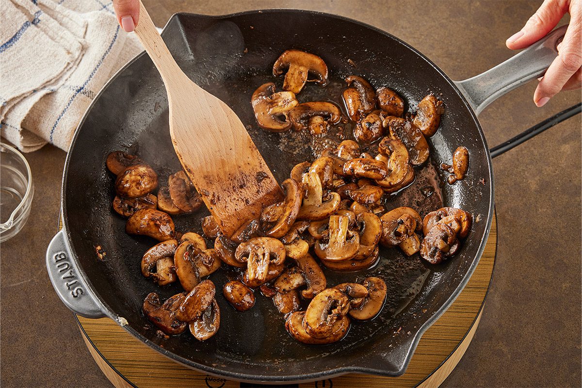 3/4 angle view shot of a Sliced mushrooms are being sautéed in a black skillet and stirred by a hand with a wooden spatula and A towel and glass bowl sit nearby;