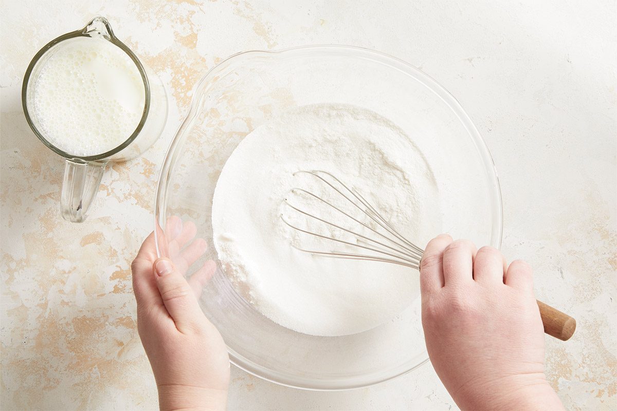 Overhead shot of a person whisking flour and sugar in a clear glass bowl, with a pitcher of milk beside it;