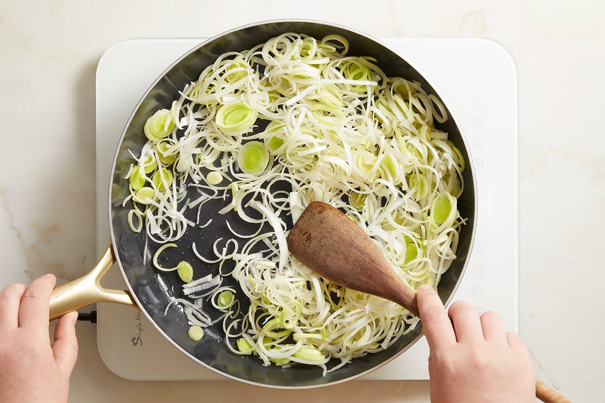 Overhead shot of sliced leeks sautéing in a skillet, as hands stir the mixture with a wooden spoon over a white cutting board.