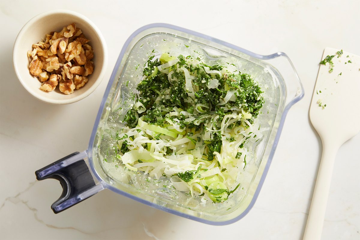 Overhead shot of shredded vegetables and herbs combined in a blender jar, with a small bowl of chopped nuts resting nearby on the counter.