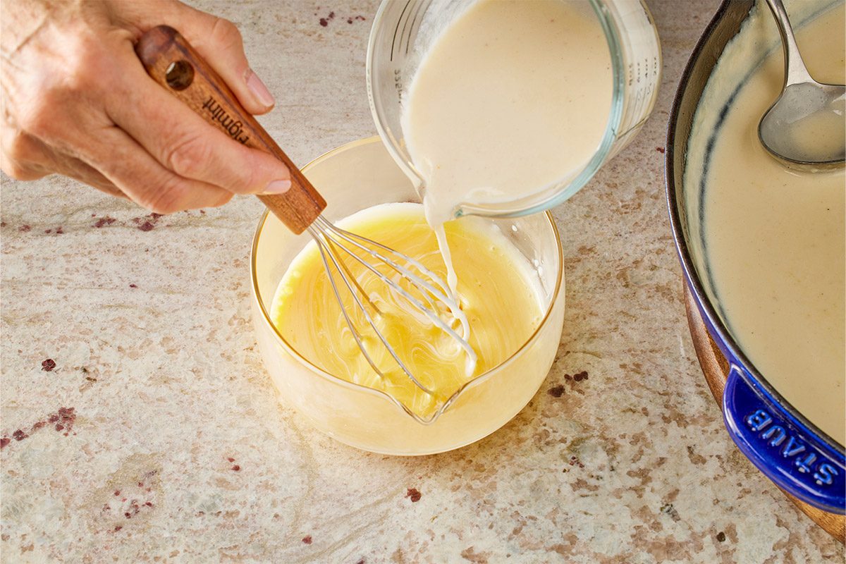 Overhead shot of a speckled countertop two hands are visible one whisking beaten eggs in a bowl while the other pours a creamy mixture from a glass measuring cup into it nearby sits a blue pot containing sauce with its lid and metal spoon resting close by all kitchen utensils and surfaces show smooth clean textures