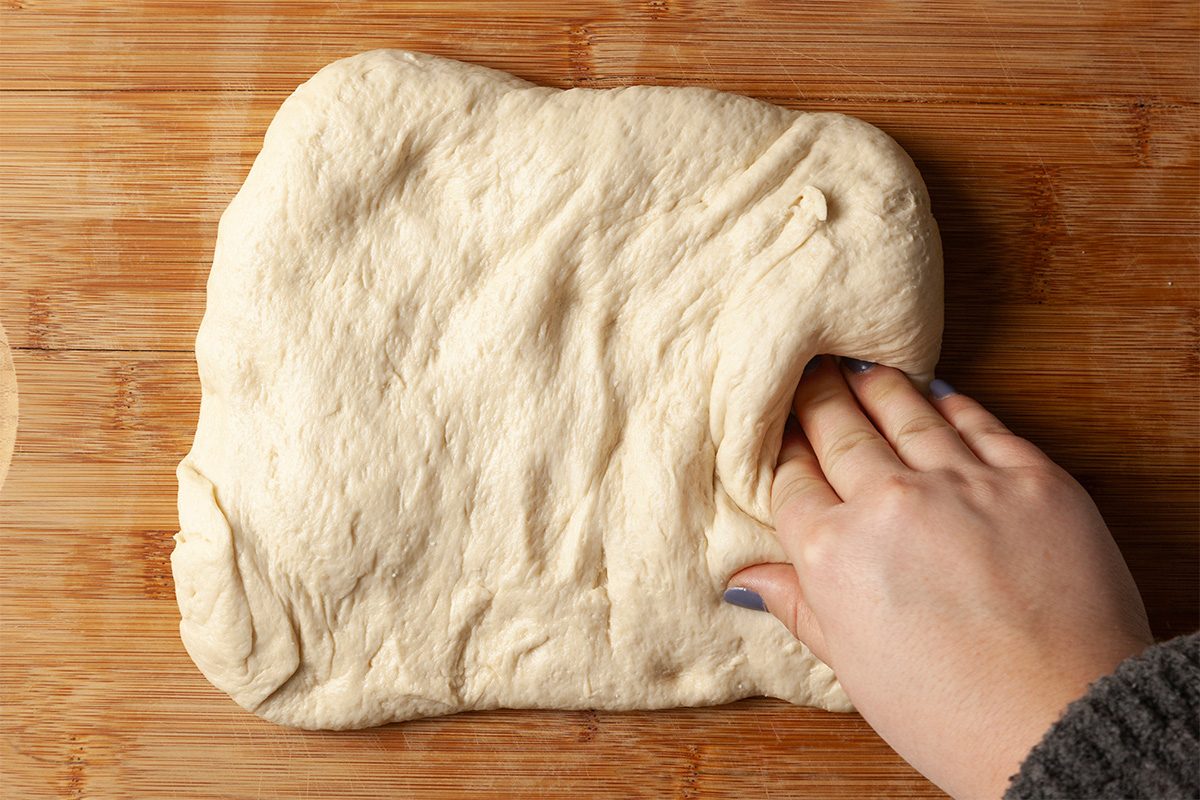 Overhead shot of a hand stretching and pressing soft pizza dough on a wooden board, showing the texture and elasticity of the dough as it’s shaped;